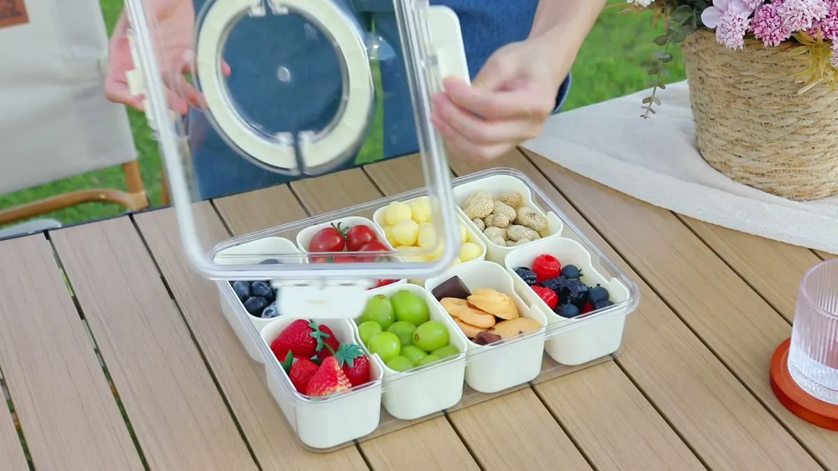 Se abre la tapa del Snackle Box en una mesa del jardín: frutos rojos, uvas, fruta y frutos secos en sus compartimentos
